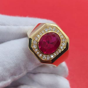 May include: A gold ring featuring a large, oval-cut ruby center stone. The ruby is surrounded by a halo of small, round diamonds. The ring has a geometric design with a polished finish.