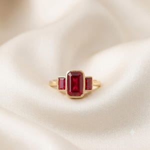 May include: A gold ring featuring a large emerald-cut red ruby center stone flanked by two smaller emerald-cut red ruby side stones. The three rubies are set in a bezel setting on a polished gold band, creating a classic three-stone design.