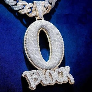 May include: A diamond-encrusted pendant necklace with the letter "O" and the word "BLOCK" in a stylized font. The pendant is attached to a thick, silver chain. The background is a deep blue.