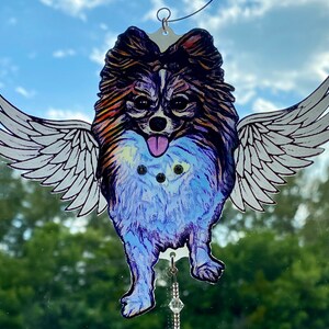 May include: A colorful, hand-painted ornament of a dog with wings. The dog is brown, black, and white with a blue and purple body. The wings are white with black outlines. The ornament has a crystal hanging from it.