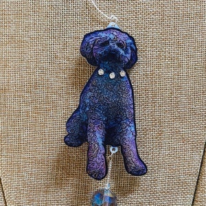 May include: A blue and purple dog ornament with a rhinestone collar hanging from a silver chain. The ornament is on a brown burlap background.