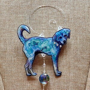 May include: A blue and teal dog ornament with a silver wire hanger. The dog has a watercolor effect, with a crystal collar and a dangling iridescent glass ball. The ornament is designed to hang.