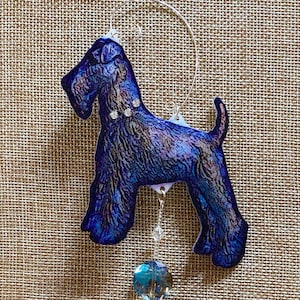 May include: A blue and purple iridescent glass ornament of a dog with a crystal dangle. The dog is a wirehaired terrier breed.