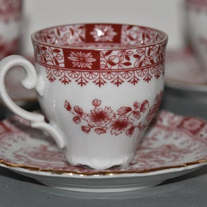 Può includere: Una tazza da tè bianca con un motivo floreale rosso e bianco. La tazza da tè ha un manico e si trova su un piattino abbinato.
