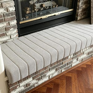 May include: A light gray, textured fabric cushion cover featuring a series of vertical, padded channels. The cover is designed to fit over a rectangular bench or hearth surface, showcasing a consistent, repeating ribbed pattern across its length.