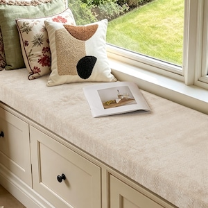 May include: A window seat with a beige herringbone cushion, featuring three decorative pillows: one solid green, one floral print, and one cream with abstract brown and black shapes. An open book with a food photograph rests on the cushion.