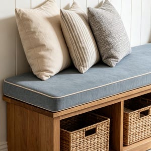 May include: A wooden bench with a blue fabric cushion featuring white piping. Three square throw pillows are arranged on top: one solid beige, one with vertical brown and white stripes, and one textured grey. Two woven wicker baskets sit below.
