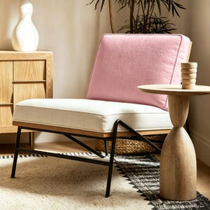 May include: An accent chair featuring a soft pink upholstered backrest and a cream-colored seat cushion. The chair is supported by a light wood frame and thin black metal legs. Beside it stands a light wood side table with a cylindrical base.