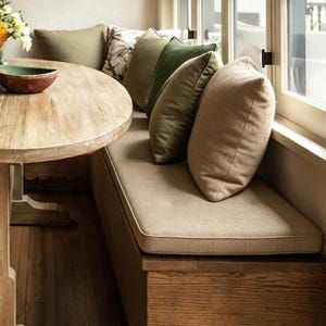 Könnte beinhalten: Eine hölzerne Sitzecke mit einer langen Bank, einem beigen Sitzpolster und mehreren Zierkissen in Olivgrün und Beige. Links steht ein runder Holztisch, auf dessen Oberfläche nahe dem Rand eine dunkelbraune Schale platziert ist.