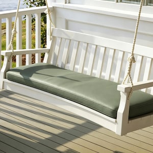 Op de afbeelding: Een witte houten veranda-schommelbank met een lattenrugleuning en armleuningen, opgehangen aan dikke gedraaide touwen. De zitting is voorzien van een lang, rechthoekig, saliegroen kussen dat de gehele lengte van de bank beslaat.