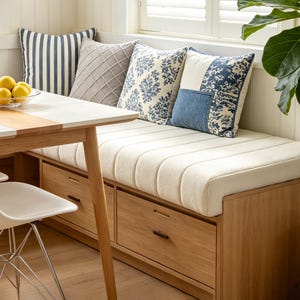 May include: A wooden bench with two storage drawers and a cream-colored tufted cushion. Four decorative throw pillows are arranged on top: one with blue stripes, one with a grey grid pattern, and two with blue and white floral and denim designs.