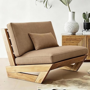 May include: A low-profile lounge chair featuring a natural light-toned wooden frame with angled supports. The chair includes a thick, textured tan seat cushion, a matching backrest cushion, and a small rectangular lumbar pillow in the same color.