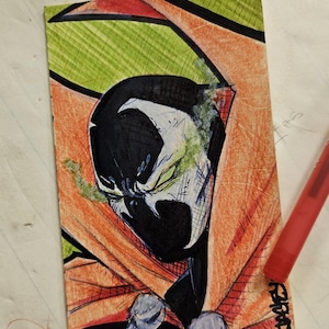 May include: Hand-drawn illustration of Spawn, a comic book character, with a black and white face, a red cape, and green and orange background. The artwork is signed in the bottom right corner.