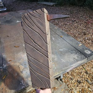 May include: A brown, rectangular anvil with a textured, wood-grain design on one side. A copper-coloured horn extends from the top. The anvil is held by a person. The anvil is a tool used for shaping metal.