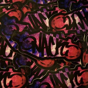 May include: A stack of black and white stickers with a galaxy background. The stickers feature the word "SNACKS" in a graffiti style font. The galaxy background is a mix of pink, purple, and blue.