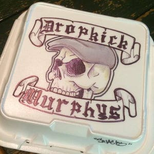 May include: A white styrofoam container with a hand-drawn design of a skull wearing a cap and a banner that reads "Dropkick Murphys".