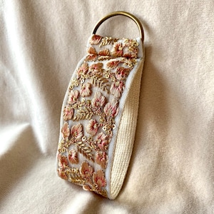 Op de afbeelding: Een witte sleutelhanger van stof met een gouden metalen ring. De stof is geborduurd met roze en gouden bloemenmotieven.