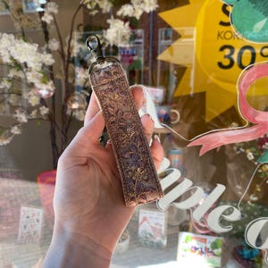 May include: A rectangular fabric keychain featuring an intricate floral embroidery pattern in shades of mauve, purple, and gold. The strap is finished with a bronze-toned metal lobster clasp at the top, providing a secure attachment point for keys.