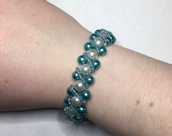 May include: A beaded bracelet with alternating turquoise and white beads. The bracelet is woven in a zig-zag pattern.