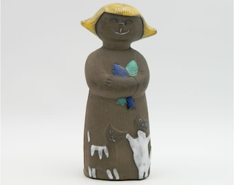 Könnte beinhalten: Keramikfigur einer lächelnden Person mit gelbem Haar, die blaue und grüne Objekte hält. Die Figur ist braun mit weißen Akzenten und einem Katzendesign. Die Figur ist ein dekorativer Artikel.