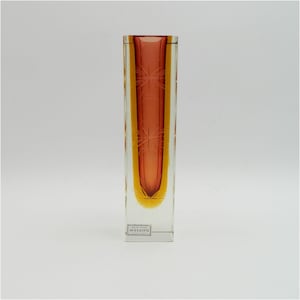 Könnte beinhalten: Eine hohe, klare Glasvase mit einem mehrschichtigen Design in Rot und Gelb. Die Vase hat einen quadratischen Boden und ist mit einem Sternmuster graviert. Die Vase ist mit "Murano" beschriftet.