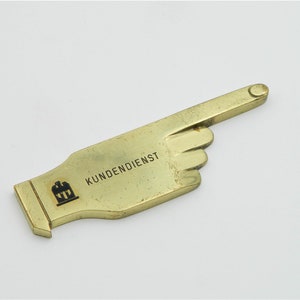 Könnte beinhalten: Eine goldfarbene Metallhand, die nach rechts zeigt, mit dem Wort "KUNDENDIENST" und einem kleinen schwarz-weißen Logo auf der Handfläche.