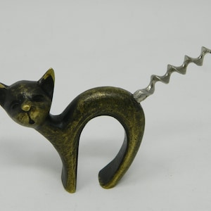 Puede incluir: Un sacacorchos con forma de gato de color bronce. El gato está en una pose juguetona con un cuerpo curvado y una cola de sacacorchos plateada. Las orejas y la cara del gato tienen un diseño detallado.