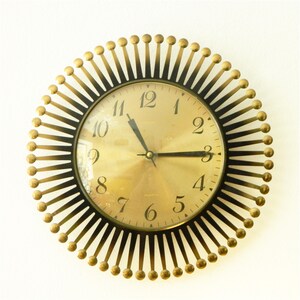 Könnte beinhalten: Eine runde Wanduhr mit goldfarbenem Zifferblatt und schwarzen Ziffern. Die Uhr hat ein Strahlen-Design mit strahlenförmig angeordneten schwarzen Metallstäben, die jeweils mit einer kleinen goldenen Kugel versehen sind. Schwarze Zeiger in der Mitte.