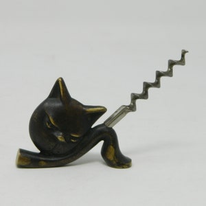 Puede incluir: Un sacacorchos de bronce con forma de gato, con una espiral de metal plateado. El gato está enroscado con la cola envuelta alrededor de su cuerpo.