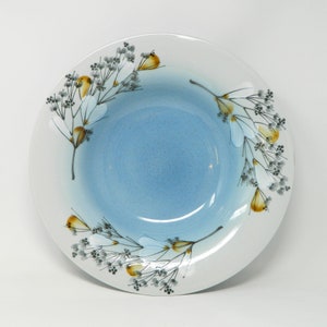 Può includere: Una ciotola in ceramica bianca con una glassa blu e un motivo floreale. Il motivo presenta fiori marroni e gialli con steli e foglie grigi.