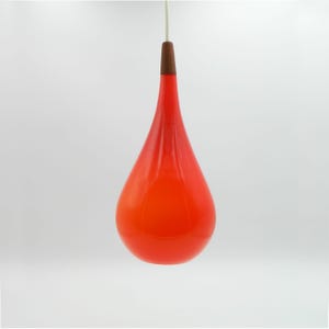 Peut inclure: Suspension luminaire en verre rouge en forme de goutte d'eau. Le luminaire est doté d'un sommet en bois et d'un cordon blanc. Il est suspendu sur un fond blanc. La couleur rouge vif est un élément marquant.