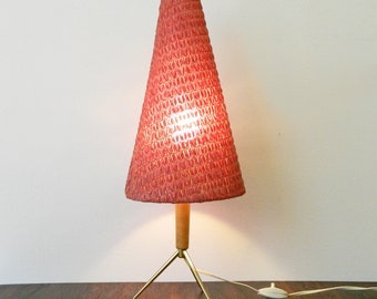 May include: A red cone-shaped lampshade with a wooden base and three gold metal legs. The lamp is turned on and the light is shining through the lampshade.