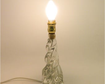 May include: A clear glass table lamp with a twisted, textured base. The lamp has a gold-colored bulb socket and a lit, white, teardrop-shaped bulb. A white electrical cord extends from the base. The lamp is approximately 10 inches tall.