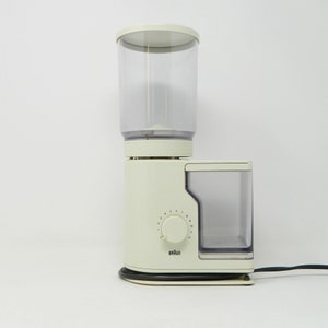 Könnte beinhalten: Eine weiße Braun Kaffeemühle mit einem transparenten Kunststoffbehälter für Kaffeebohnen und einem transparenten Kunststoffbehälter für gemahlenen Kaffee. Die Mühle hat ein Zifferblatt mit den Zahlen 1 bis 12 zur Auswahl der Mahlgradgröße.