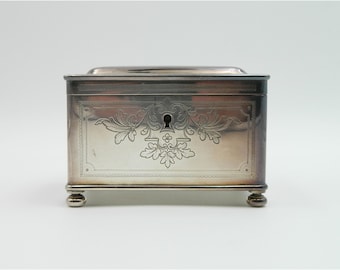 May include: A silver rectangular box with a decorative floral design and a keyhole on the front. The box has four small feet.