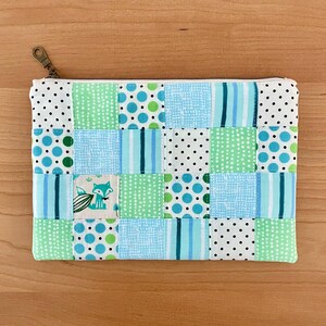 Könnte beinhalten: Ein Patchwork-Etui mit Reißverschluss, das Quadrate aus blauem, grünem und weißem Stoff mit Punkten, Streifen und einem Fuchsmotiv aufweist. Das Etui hat einen messingfarbenen Reißverschluss und ist für kleine Gegenstände gedacht.