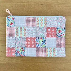 May include: A patchwork zippered pouch in shades of pink, white, and blue. The rectangular pouch features a variety of fabric patterns, including floral, gingham, and wave designs. A rose gold zipper and pull complete the design.