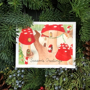 May include: A watercolor Christmas card featuring mushroom houses with red caps and brown stems. The card has the text "Season's Greetings" and is surrounded by evergreen branches and pine cones. A small bird is carrying a garland.