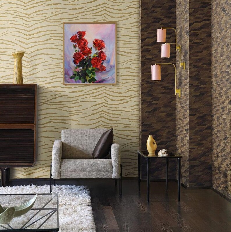 May include: An interior scene featuring a framed painting of red roses, a gray armchair with a brown pillow, and a black side table. The walls have patterned wallpaper, and the floor is dark wood. Decorative lamps with pink shades are mounted on the wall.