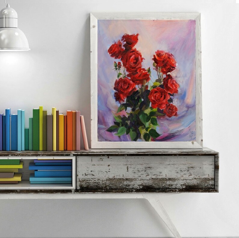 May include: An oil painting in a white frame depicts a bouquet of vibrant red roses with green leaves against a soft, colorful background. The artwork is displayed on a distressed, white wooden shelf, with a collection of colorful books to the left.