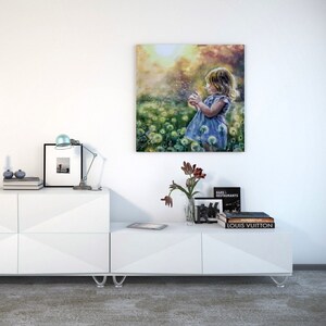 May include: A framed canvas art print shows a young child in a blue dress blowing dandelion seeds in a field of flowers. The artwork is displayed on a white wall above a white modern media unit with a lamp, books, and decorative items.