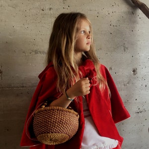 May include: A young person dressed in a Little Red Riding Hood costume, featuring a vibrant red cape and skirt. The outfit includes a white top with a ruffled hem and a woven basket. The background is a textured gray wall.