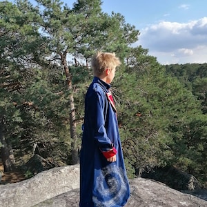 May include: A long, navy blue coat with red and white detailing. The coat has a distinctive spiral pattern on the lower part. The person is standing on a rock formation with a forest backdrop.