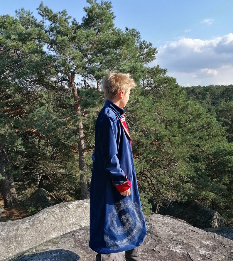 May include: A long, navy blue coat with red and white detailing. The coat has a distinctive spiral pattern on the lower part. The person is standing on a rock formation with a forest backdrop.