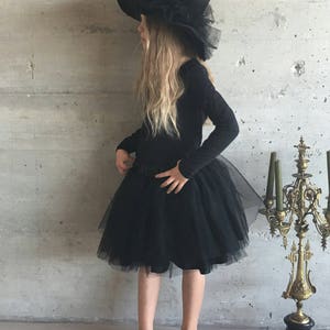 May include: A young girl dressed as a witch for Halloween. She is wearing a black tulle skirt, a black long-sleeved shirt, and a large black witch hat with a tulle trim.