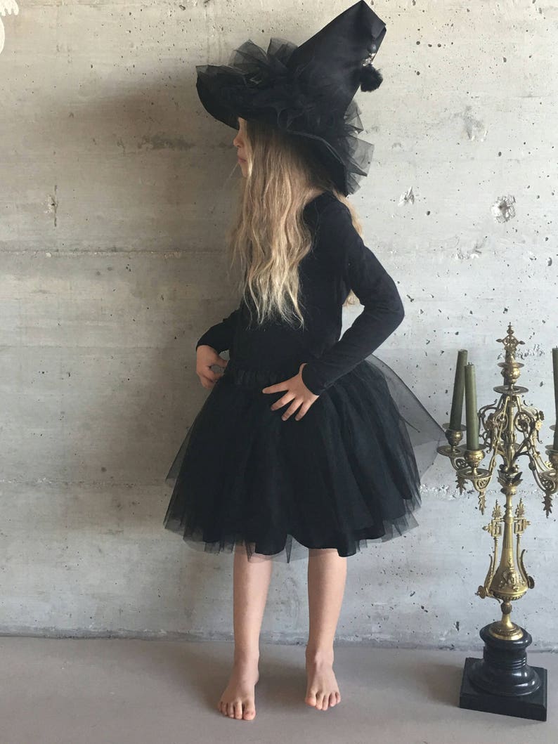 May include: A young girl dressed as a witch for Halloween. She is wearing a black tulle skirt, a black long-sleeved shirt, and a large black witch hat with a tulle trim.