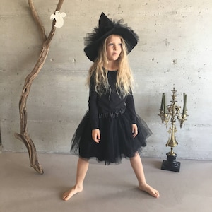 May include: A young girl dressed as a witch for Halloween. She is wearing a black tulle skirt, a black long-sleeved shirt, and a black witch hat with a tulle trim.