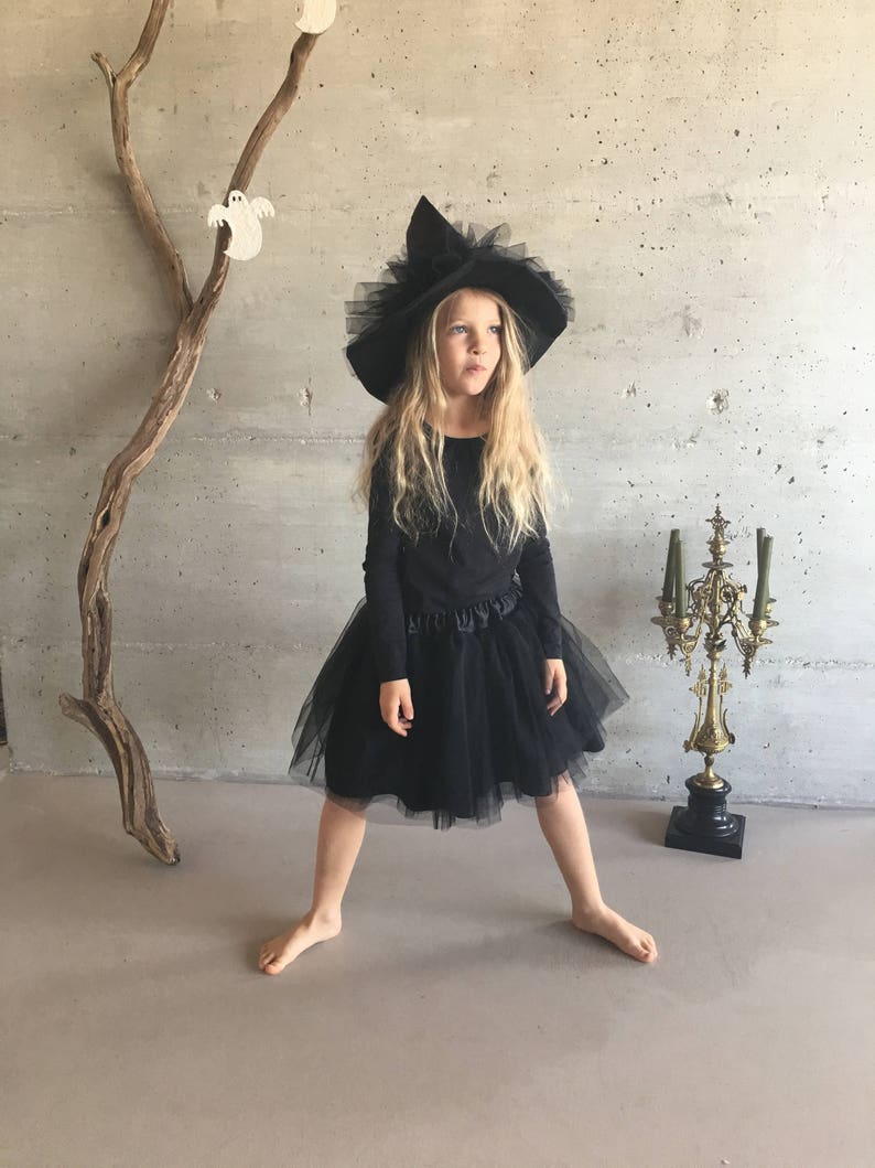 May include: A young girl dressed as a witch for Halloween. She is wearing a black tulle skirt, a black long-sleeved shirt, and a black witch hat with a tulle trim.