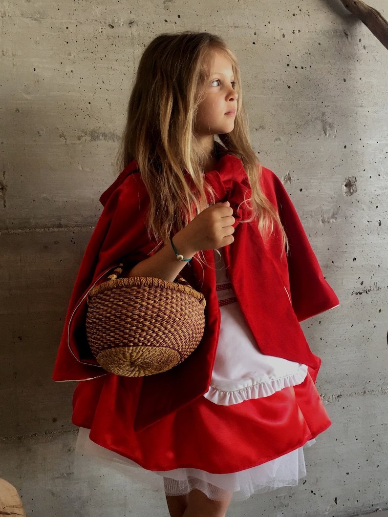 May include: A young person dressed in a Little Red Riding Hood costume, featuring a vibrant red cape and skirt. The outfit includes a white top with a ruffled hem and a woven basket. The background is a textured gray wall.