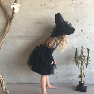 May include: A young girl dressed as a witch for Halloween. She is wearing a black tulle skirt and a black witch hat with a wide brim and a black feather trim. She is standing in front of a gray wall with a wooden branch behind her.